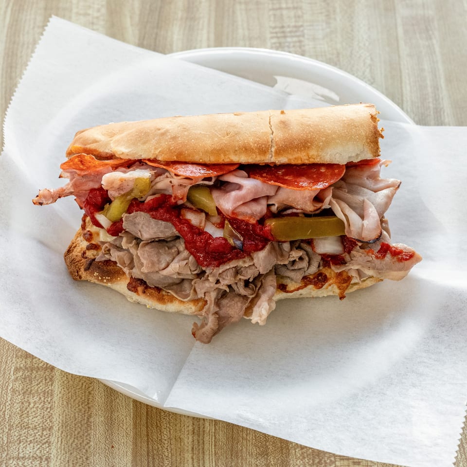 A large Raydee Magoo's sub sandwich, loaded with meats, sauce, and pickles, on a white plate.