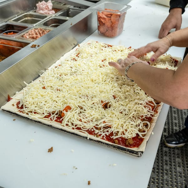 Hands spreading shredded mozzarella cheese over a large rectangular pizza with sauce and toppings.