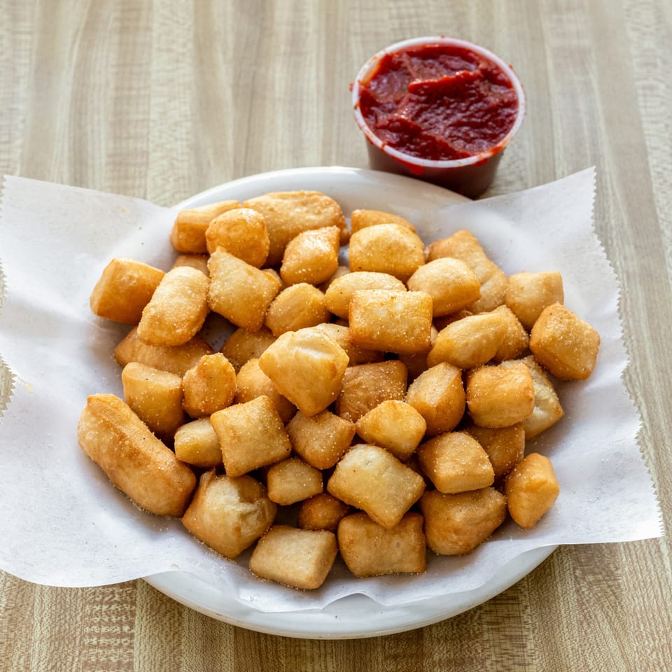 Golden-brown fried dough knots, sprinkled with seasoning, served with a side of red marinara sauce.