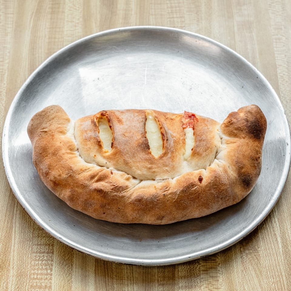 A golden-brown calzone with visible cheese and a hint of sauce, served on a metal pizza pan.