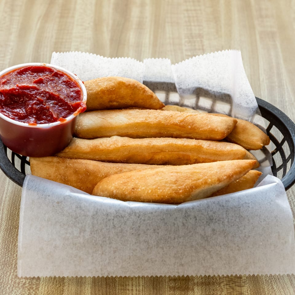 Golden-brown breadsticks with a side of red marinara sauce in a black basket lined with white paper.
