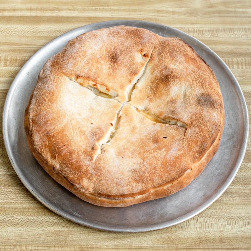 A golden-brown, baked calzone with a cross-shaped cut on top, served on a metal pizza pan.